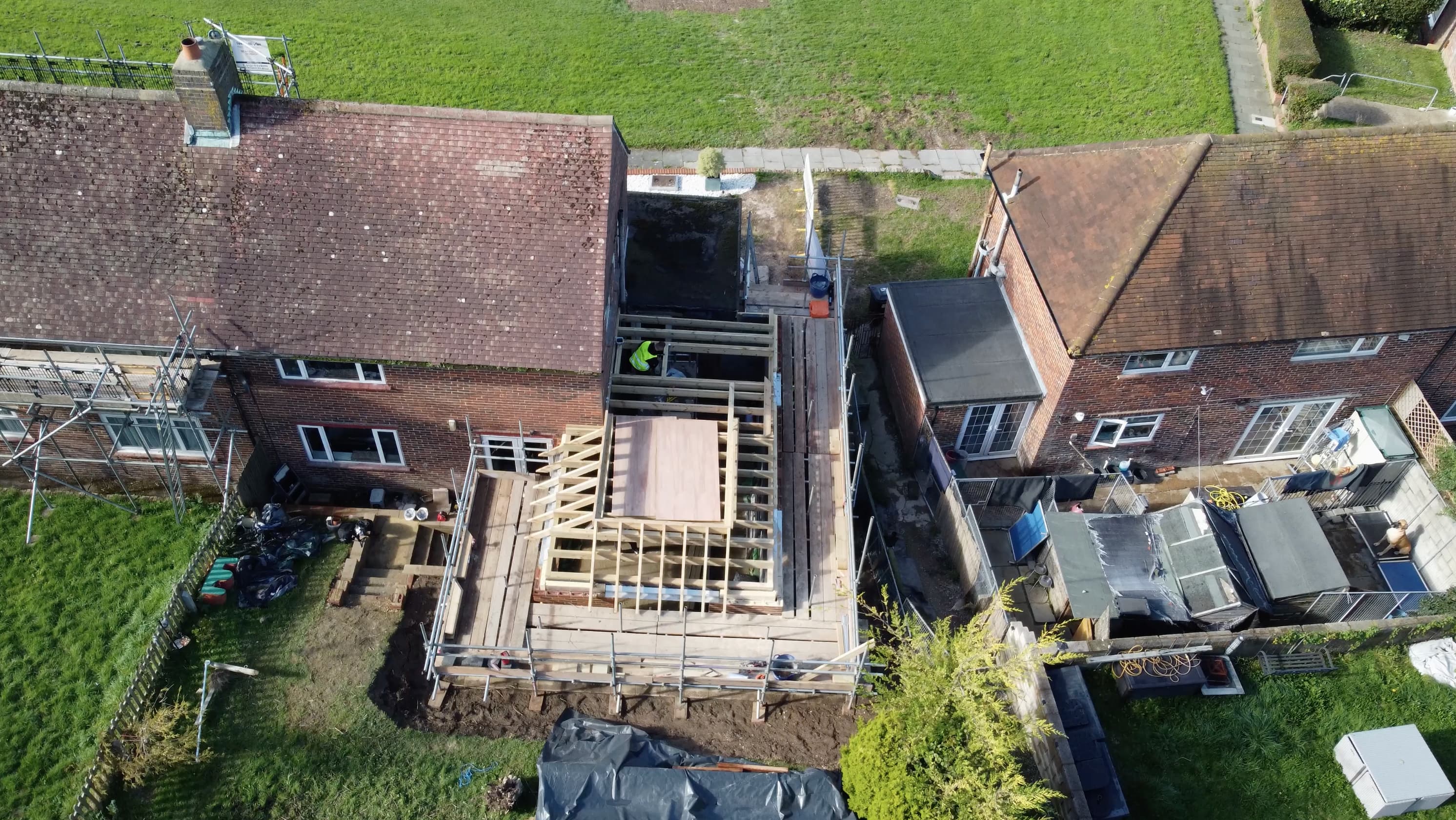 Drone photograph looking down on a brick semi detached house with a large rear extension under construction: timber roof frame, scaffolding, a worker in a hi vis vest, and neighbouring gardens on either side.